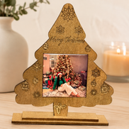 A gold-toned laser-cut Christmas tree picture frame with engraved holiday motifs and 'Merry Christmas' text, displayed on a wooden surface. 