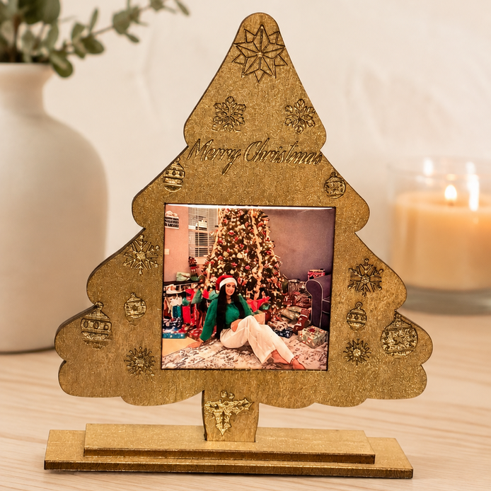 A gold-toned laser-cut Christmas tree picture frame with engraved holiday motifs and 'Merry Christmas' text, displayed on a wooden surface. 